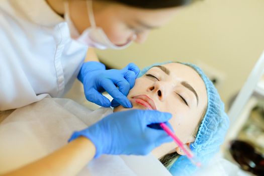 Skilled cosmetologist shaping lips with pencil for pretty girl before microblading. Concept of permanent makeup artist and beauty salon.