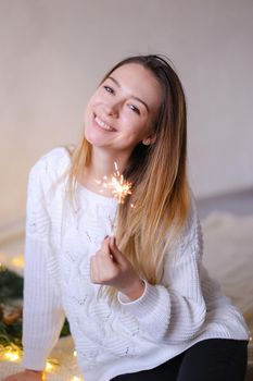 Young happy woman sitting with bengal light near twinkling yellow garlands. Concept of positive emotions and celebration New Year holidays.