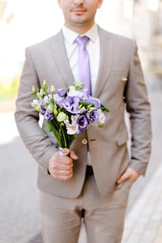Young caucasian groom keeping bouquet of flowers and wearing grey suit. Concept of wedding photo session and waiting for bride.