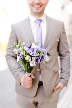 Young caucasian groom keeping bouquet of flowers and wearing grey suit. Concept of wedding photo session and waiting for bride.