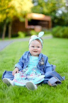 Little happy female baby sitting on green grass and wearing jeans jacket. Concept of childhood and resting in garden, nature background.