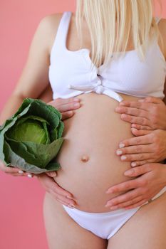 Closeup pregnant woman in underwear keeping cabbage in pink monophonic background. Concept of expactant photo session.