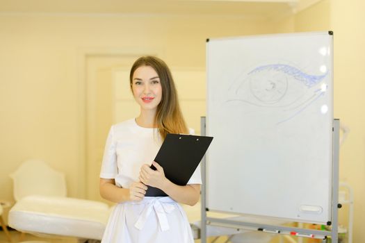 Young cosmetologist standing with black folder near placard with drawn eye at cosmetology cabinet. Concept of qualitative medical worker, beauty salon and permanent makeup.