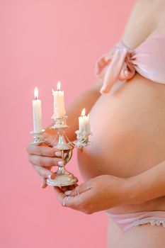 Closeup happy pregnant woman in underwear holding belly and keeping candles in pink monophonic background. Concept of expactant photo session.
