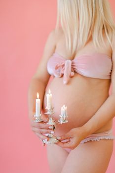 Blonde happy young pregnant woman in underwear holding belly and keeping candles in pink monophonic background. Concept of expactant photo session.