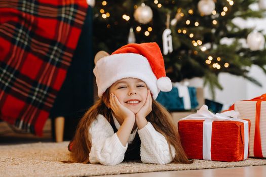 A happy little girl in a Santa Claus hat smiles with gifts in her hands.