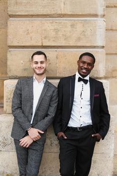 Afro american and caucasian smiling gays standing near building and wearing suits. Concept of lgbt and walking in city.