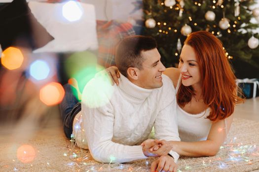 a happy married couple is lying on the floor near the Christmas tree at home.