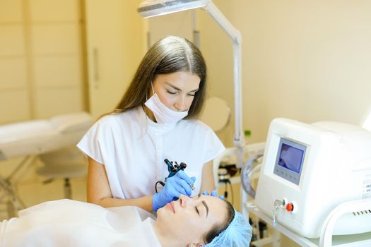 Caucasian cosmetologist making permanent makeup for girl at beauty salon. Concept of cosmetology equipment and microblading.
