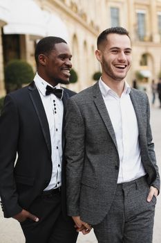 Afro american and caucasian happy handsome gays walking outside and holding hands in city. Concept of same sex male couple.