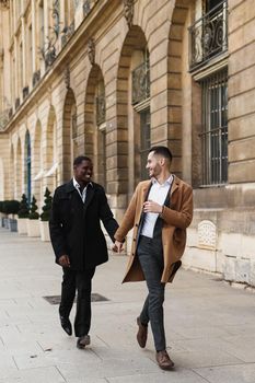 Caucasian smiling man in suit walking with afroamerican male person and hugging in city. Concept of happy lgbt gay and strolling.