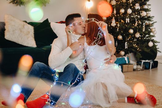 a happy married couple is sitting at home on the floor near the Christmas tree and kissing.