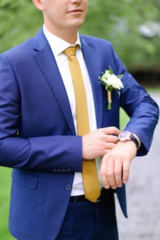 Young caucasian groom looking at watch and wearing blue jacket with flower. Concept of wedding photo session and waiting for bride.