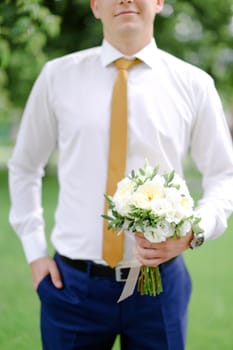 Young caucasian groom keeping bouquet of flowers and wearing white shirt. Concept of wedding photo session and waiting for bride.