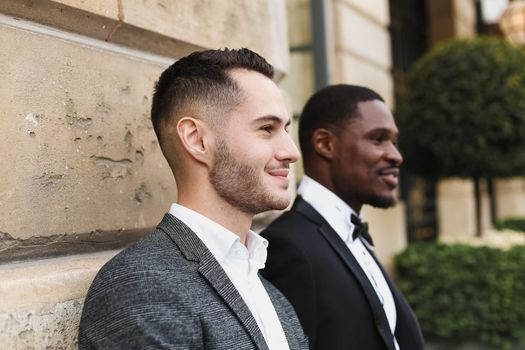 Afro american and european gays standing near building and wearing suits. Concept of lgbt and walking in city.