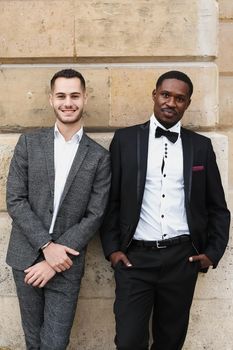 Afro american and european gays standing near building and wearing suits. Concept of lgbt and walking in city.