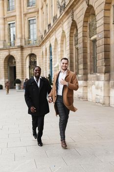 Caucasian smiling man in suit walking with afroamerican male person and hugging in city. Concept of happy lgbt gay and strolling.