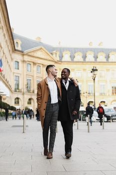 Caucasian smiling man in suit walking with afroamerican male person and hugging in city. Concept of happy lgbt gay and strolling.