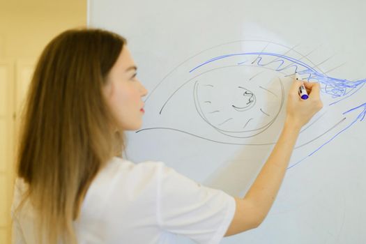 Female cosmetologist draws eye on placard at cosmetology cabinet, focus on drawing. Concept of makeup courses and beauty salon.