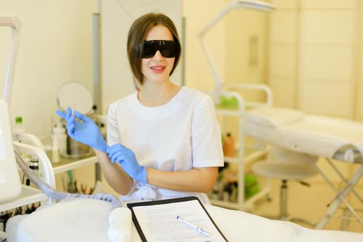 Young female cosmetologist wearing special purpose glasses and latex gloves near permanent makeup device. Concept of beauty cosmetology salon and expensive high quality equipment.