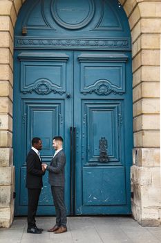 Afro american smiling man holding hands of caucasian boy in blue door background, wearing suit. Concept of same sex couple and gays.