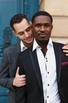 Two boys, caucasian and afro american, wearing suits standing near building and hugging. Concept of gays and lgbt.