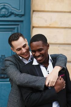 Two boys, caucasian and afro american, wearing suits standing near building and hugging. Concept of gays and lgbt.
