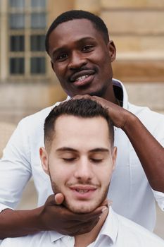 Afro american boy holding caucasian guy head by hands, wearing white shirt. Concept of stylish boy and haircut. Same sex couple, lgbt gays