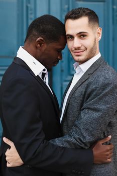 Afro american handsome man hugging caucasian guy in door background, wearing suit. Concept of same sex couple and gays.