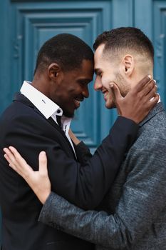 Afro american handsome man hugging caucasian guy in door background, wearing suit. Concept of same sex couple and gays.