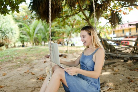 Young happy woman using laptop and riding on swing on sand, wearing jeans dress. Concept of summer vacations and modern technology.