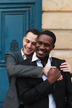 Two boys, caucasian and afro american, wearing suits standing near building and hugging. Concept of gays and lgbt.