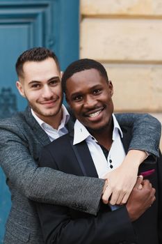 Two boys, caucasian and afro american, wearing suits standing near building and hugging. Concept of gays and lgbt.