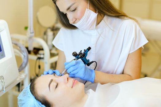 Skilled cosmetologist doing permanent makeup for girl at beauty salon. Concept of cosmetology equipment and microblading.