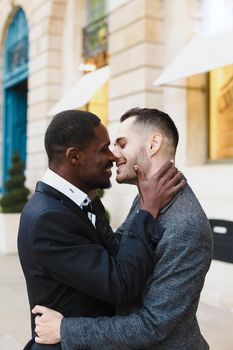 Afro american gay kissing caucasian boy outside, wearing suits. Concept of lgbt and same sex couple.