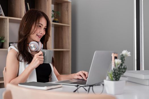 Asian woman with cup of coffee using laptop computer with headset at home, Online learning education concept.