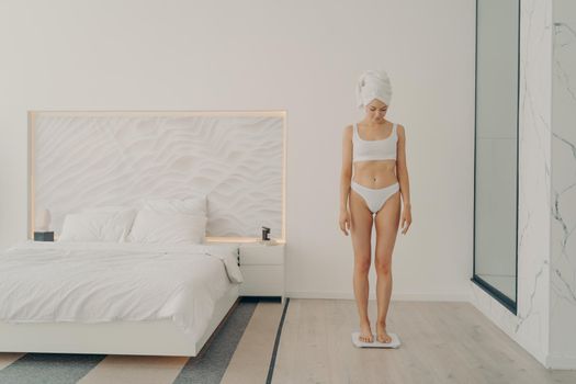Beautiful young good looking female in underwear with head wrapped in towel looking down while standing in bedroom on scale and measuring her weight. Beauty and body care concept