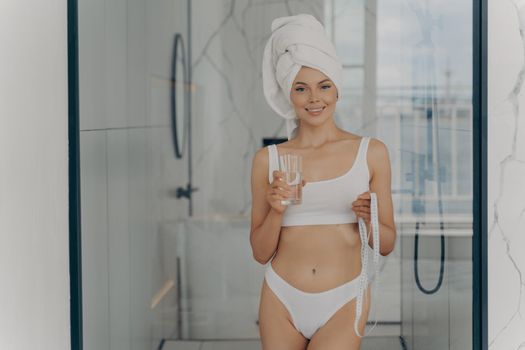 Slender happy athletic woman in white classic underwear standing in bathroom after shower routine with measuring tape in one hand and holding glass of water in other hand, healthy lifstyle concept