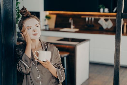 Young woman in silk pajama staying at home and enjoying morning cup of coffee while applying white skincare disposable mask on face. Daily routine procedure for rejuvenation and care of skin