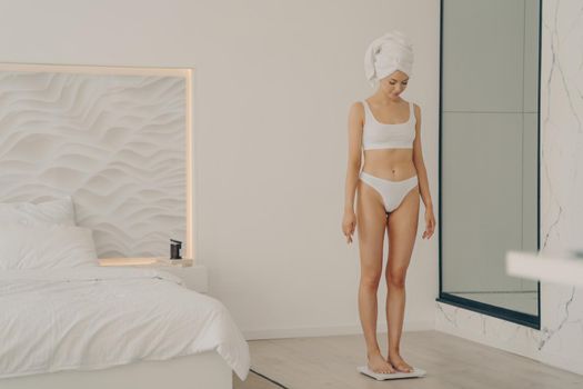 Attractive slim young girl standing on scales in bedroom after morning shower in white underwear and bath towel on head, doing daily measurement of body weight. Healthy lifestyle and dieting concept