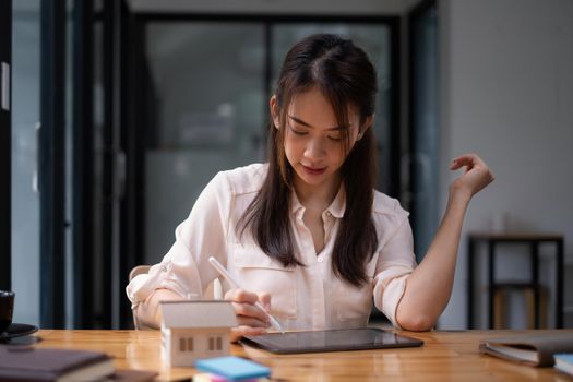 Architect woman using a digital tablet application to hand-draw a design house with a model on a wooden desk. Property idea in real estate