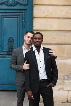 Two boys, caucasian and afro american, wearing suits standing near building and hugging. Concept of gays and lgbt.