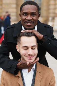 Afro american boy holding caucasian guy head by hands, wearing white shirt. Concept of stylish boy and haircut. Same sex couple, lgbt gays