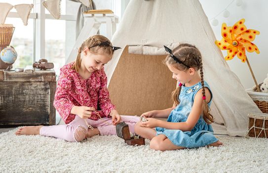 Little girls playing with small treasure box filled with jewels sitting on floor in light room on wigwam background