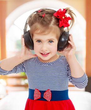 Nice little blonde girl with a red bow on her head, to listen to music through big black headphones. Close-up.On the background of the hall with a large arched window