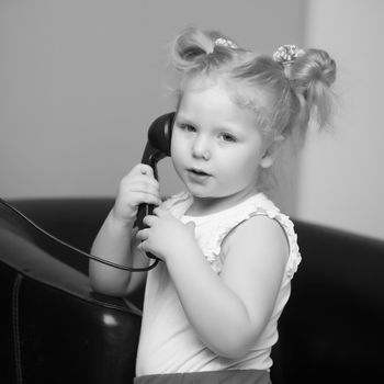 A beautiful little girl in a room in the fifties of the last century, talking on an old phone with a big black tube. Retro style, black and white photo in the studio. Concept of nostalgia, vintage.