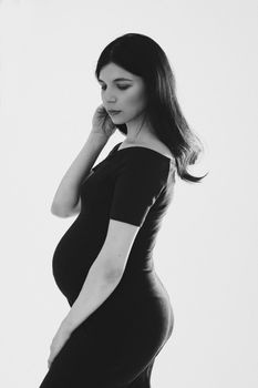 Studio black and white portrait of attractive Caucasian young woman with dark hair in black dress standing on white background, looking down. Pregnant woman with belly in tight dress.