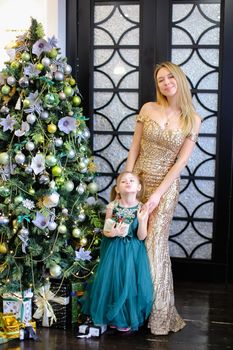 Young caucasian woman and little daughter with gift wearing dresses standing near Christmas tree. Concept of celebrating winter holidays, New Year and fashionable look.