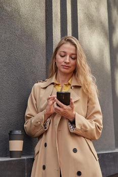 caucasian woman in beige trench coat using smartphone outdoors on sunny day, reading news, social media. attractive female street portrait at fall or spring. autumn lifestyle, modern communication