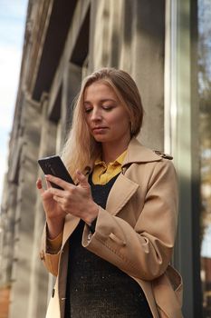 caucasian woman in beige trench coat using smartphone outdoors on sunny day, reading news, social media. attractive female street portrait at fall or spring. autumn lifestyle, modern communication